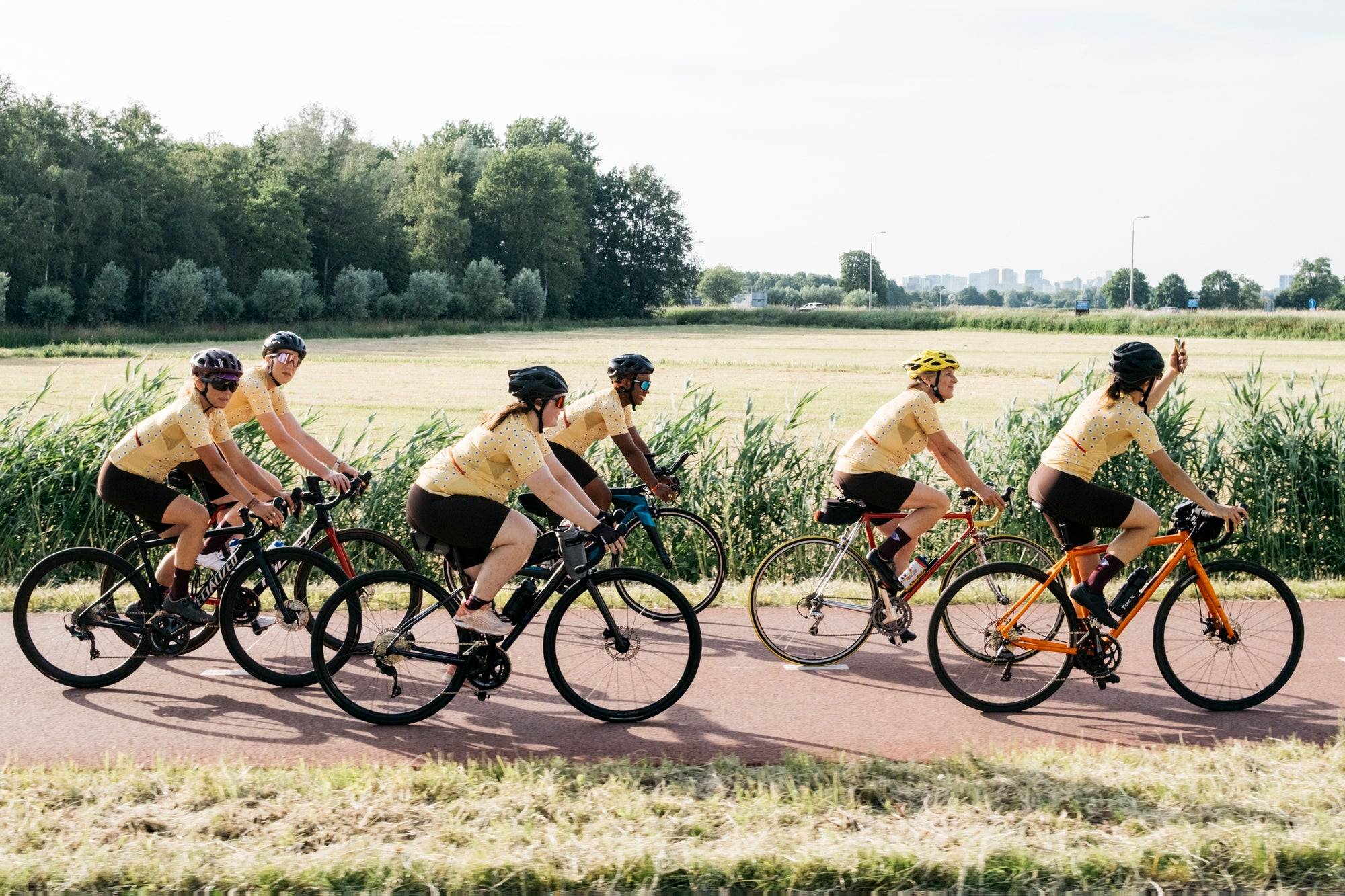 Five cyclists ride in a group along a paved path in an outdoor rural setting, wearing matching yellow jerseys and black shorts. Trees and grass are visible in the background, with one cyclist riding an orange bicycle.